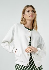 Biała bawełniana kurtka z okrągłym dekoltem, złotymi zatrzaskami, prążkowanymi mankietami i dołem. Nosi się ją na zieloną i białą paskowaną bluzkę.