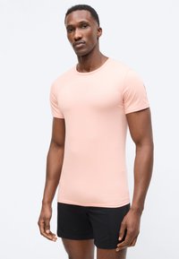 Άνδρας με κοντά μαλλιά και μεσοσκούρο δέρμα, που φοράει στενή ανοιχτό ροζ μπλούζα και μαύρο σορτς, στέκεται μπροστά σε απλό ανοιχτόχρωμο φόντο.