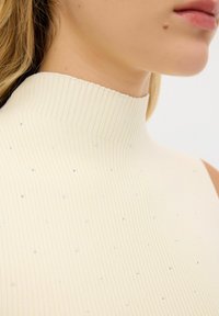 Geribbelde, mouwloze top met hoge hals in crème met verspreide zilveren versieringen, die een gestructureerde stof en naadloos ontwerp laat zien.