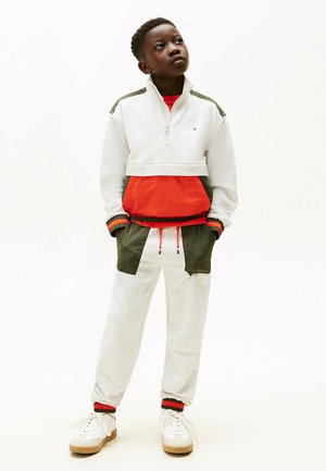 Lichtgrijze rits-sweatshirt met rode en groene accenten, gecombineerd met bijpassende grijze joggingsbroek met contrasterende groene zakken. Witte sneakers.