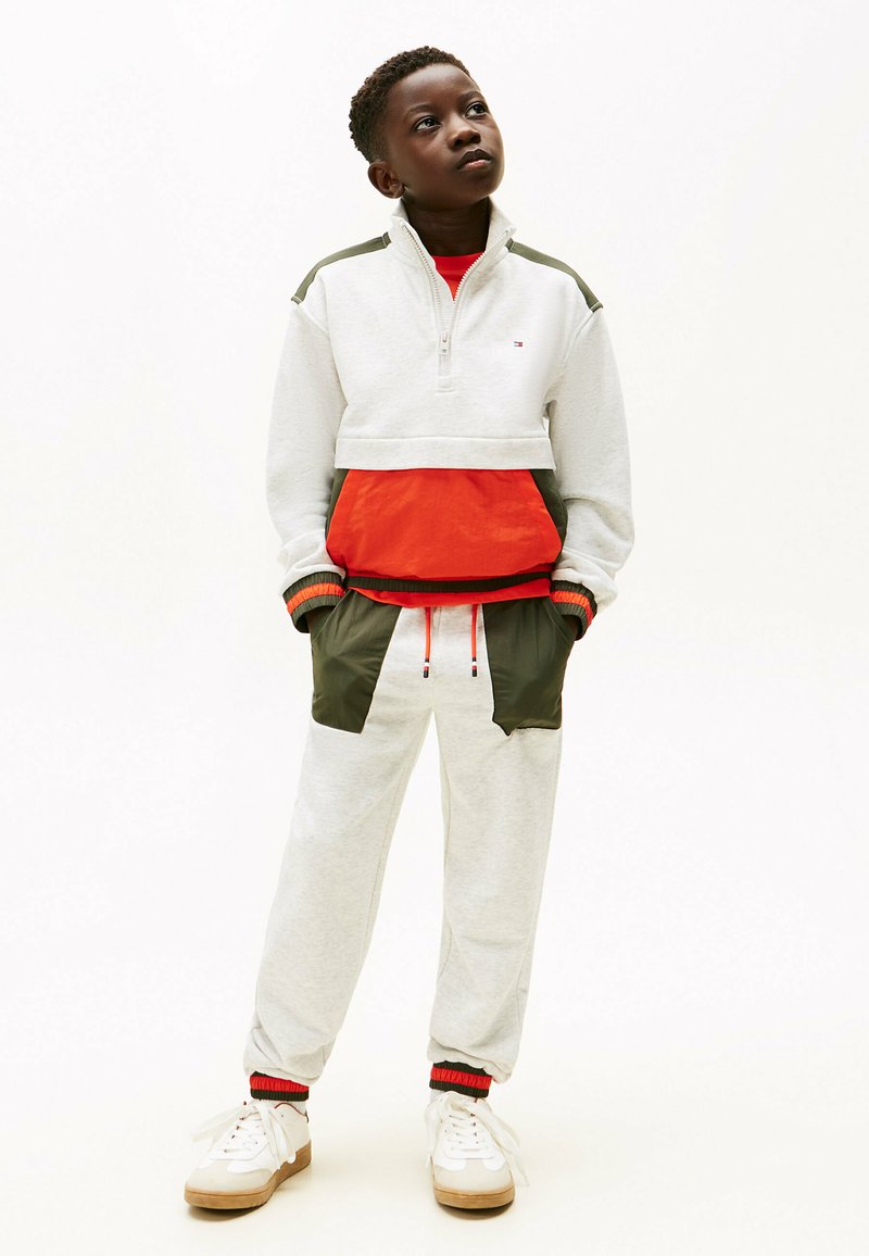 Felpa grigia chiara con zip, decorata con accenti rossi e verdi, abbinata a pantaloni sportivi grigi con tasche verdi a contrasto. Sneakers bianche.