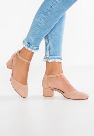 Escarpins en daim rose avec bout rond et bride à la cheville. Design à talon carré, texture lisse, associés à un jean bleu à ourlet effiloché.