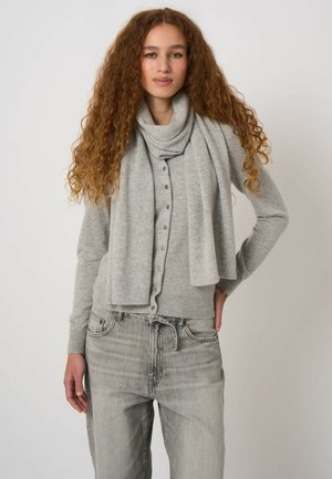 Jonge vrouw met lang krullend haar, gekleed in een lichtgrijs vest met knopen, bijpassende sjaal en vervaagde grijze spijkerbroek, staand tegen een effen achtergrond.