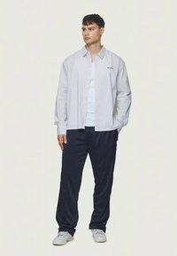 Randig långärmad skjorta i beige med knäppning fram, ihop med mörkblå byxor. Visad logotypdetalj på skjortan.