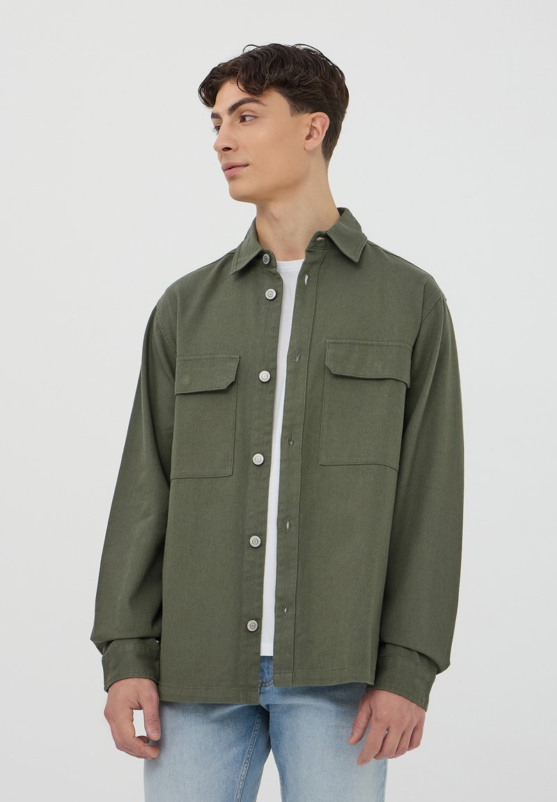 Camicia verde con bottoni in un tessuto morbido, dotata di due tasche sul petto, bottoni in metallo, maniche lunghe e una vestibilità rilassata. Indossata sopra una maglietta bianca.