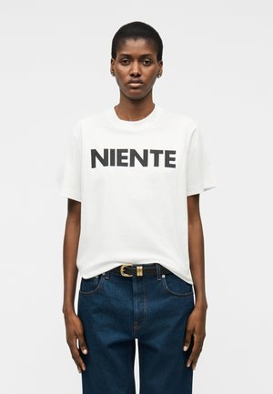Kvinne med kort hår iført en hvit T-skjorte med teksten "NIENTE", stukket inn i blå jeans med svart belte, stående mot en hvit bakgrunn.