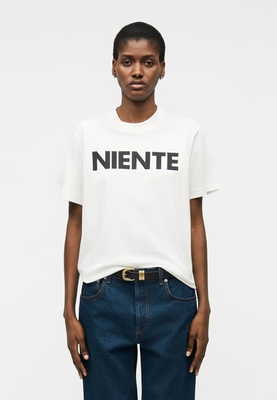 Γυναίκα με κοντά μαλλιά που φοράει άσπρο T-shirt με το κείμενο "NIENTE", μαζεμένο μέσα σε μπλε τζιν με μαύρη ζώνη, στέκεται μπροστά σε λευκό φόντο.