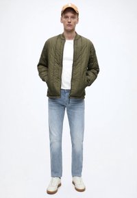 Olijfgroene doorgestepte bomberjack, wit T-shirt, lichtblauwe spijkerbroek en witte sneakers met bruine zolen. Model draagt een oranje pet.