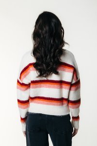 Person with long dark wavy hair wearing a white sweater with red, orange, and pink stripes, facing away against a plain background.