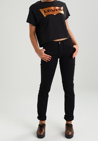 Mustat lyhyet topit, joissa on metallinhohtoinen kuparinvärinen Levi's-logo, yhdistettynä slim-fit mustiin farkkuihin ja ruskeisiin nauhakenkiin.