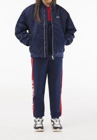 Marineblauer Bomberjacke mit Reißverschluss, gerippten Bündchen und kleinem Logo. Kombiniert mit Marine- und rot gestreiften Jogginghosen mit seitlichem Branding. Weiße Sneakers.