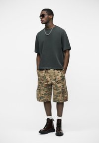 Άνδρας με μαύρα γυαλιά ηλίου, σκούρο φαρδύ μπλουζάκι, παρκετέ με παραλλαγή, άσπρες κάλτσες και μαύρα αρβυλάκια, στέκεται με τα χέρια στις τσέπες.