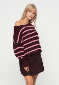 Pull rayé bourgogne et rose avec un design décalé sur les épaules. Texture tricotée et manches surdimensionnées. Porté par-dessus une robe assortie.