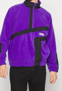 Lilla fleece-pullover med sorte detaljer og en halv-lynlås krave. Har et diagonalt design samt elastiske manchetter og kant. Lille logo-detalje.
