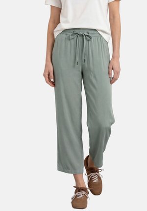 Person iført lysegrønne snøre-bukser, hvid t-shirt og brune snørebånd casual sko, stående mod en ensfarvet baggrund.