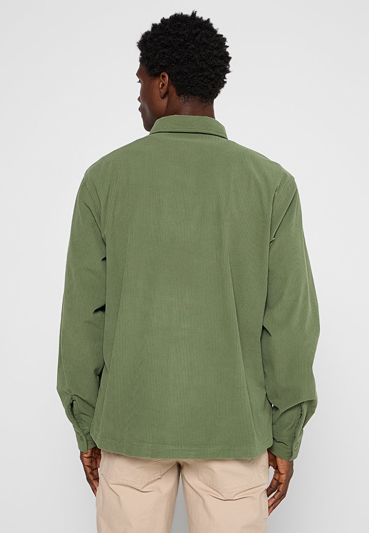 Zielona koszula z sztruksu z kołnierzykiem, długimi rękawami i mankietami zapinanymi na guziki. Tkanina ma prążkowaną fakturę i jest uzupełniona prostym krojem.
