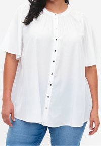 Blouse blanche à manches courtes avec boutons et texture subtilement ponctuée, portée avec un jean bleu clair, visible du cou aux hanches sur une personne.