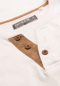 Beżowa koszula bawełniana z brązowym zamszowym paskiem na guziki i dwoma brązowymi guzikami, z okrągłym dekoltem i metką po boku.