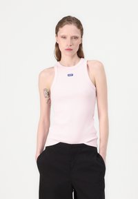 Top de malha canelada rosa clara com decote redondo e alças largas, apresentando um pequeno logotipo azul. Usado com calças pretas.