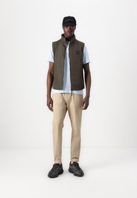 Olive green sleeveless vest over a blue t-shirt, beige trousers, and black trainers with textured soles, standing against a white background.
