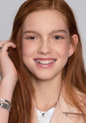 Portret van een persoon met lang, steil, roestbruin haar, gekleed in een lichtroze jasje, een zilveren ketting en een armband; lachend.