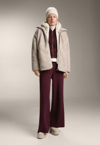 Osoba v béžovej fleecovej bunde, vínových širokých nohaviciach, bielom roláku a béžovej čiapke stojí na neutrálne pozadí.