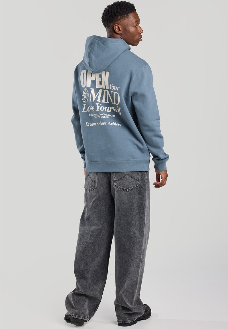 Blue hoodie with a front pocket and white text graphic on back; paired with loose-fitting gray jeans and black shoes.