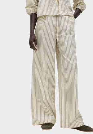 Osoba ubrana w kremowe spodnie z szerokimi nogawkami z pionowymi niebieskimi paskami, zestawione z pasującą bluzką i ciemnymi płaskimi butami.