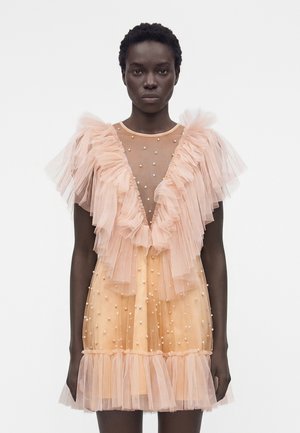 Γυναίκα που φορά διαφανές φόρεμα ροδακινί με στρωτές βολάν στους ώμους και το τελείωμα, διακοσμημένο με μικρές λευκές πέρλες, στέκεται μπροστά σε λευκό φόντο.