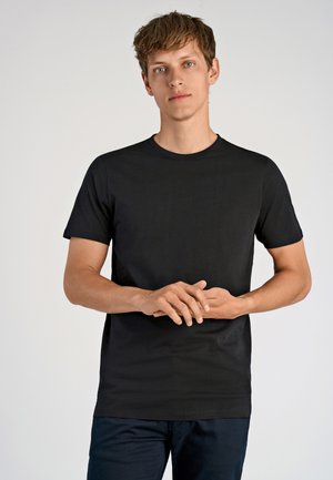 Junger Mann mit kurzem, hellbraunem Haar, der ein einfaches schwarzes T-Shirt und marineblaue Hosen trägt, steht vor einem schlichten hellgrauen Hintergrund.