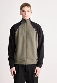 Oldalán olívazöld és fekete színblokkokkal díszített cipzáras dzseki, magas gallérral, fehér csíkozással és kis logóval a mellkason. Simább anyagú felület.