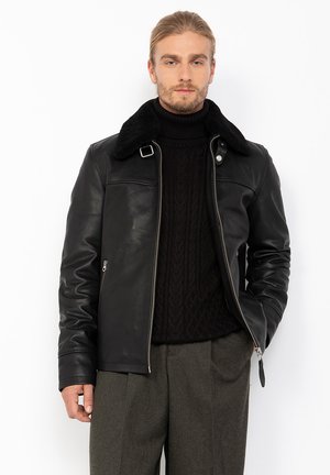 Homme portant une veste en cuir noir avec un col en shearling, un pull à col roulé en maille noire et un pantalon plié gris, se tenant devant un fond blanc.