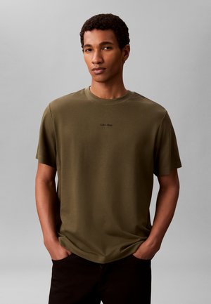 Млад мъж с къдрави къси коси, облечен в зелена тениска Calvin Klein и черен панталон на еднороден сив фон.