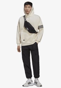 Felpa con cappuccio beige a righe nere, marsupio nero, pantaloni cargo neri e sneakers bianche con accenti neri e grigi.