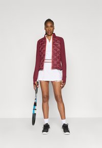 Giacca puffer bordeaux con frontale strutturato, abbinata a un polo bianco e a una gonna con bordi floreali. Inclusi scarpe da tennis nere e una racchetta.