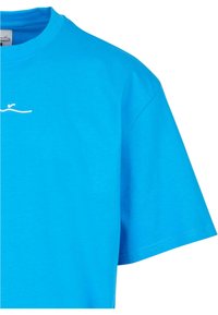 Tirkīza kokvilnas T-krekls ar apaļu kakla izgriezumu un īsām piedurknēm. Priekšpusē ir maiga balta izšūta detaļa.