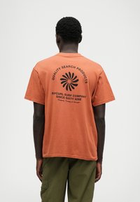 Tricou din bumbac portocaliu cu text negru și un grafic circular pe spate. Manșete scurte, guler rotund și croială lejeră.