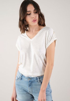 Top à manches courtes en V en tissu beige clair avec une finition texturée. Détails notables des manches retroussées. Associé à un short en jean bleu clair.