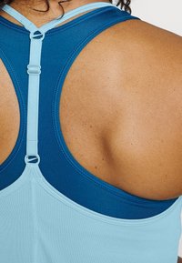 Top sportivo blu e azzurro chiaro con design a schiena scoperta, dotato di spalline regolabili e una texture liscia, che mette in evidenza la flessibilità del tessuto.