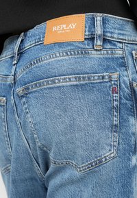 Zbliżenie tylnej kieszeni niebieskich dżinsów z brązową metką "REPLAY Since 1981" i czerwoną haftowaną literą "R" na krawędzi kieszeni.