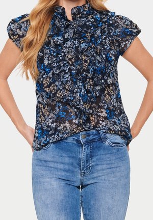 Mujer con blusa floral translúcida de manga corta con volantes y vaqueros azules, manos apoyadas en las caderas, vista desde el cuello hasta las caderas.