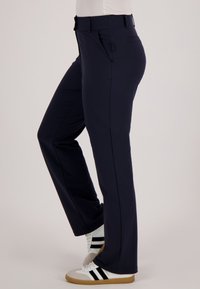 Persoon draagt donkerblauwe, rechte broekspijpen en witte sneakers met zwarte strepen, staand tegen een effen achtergrond.