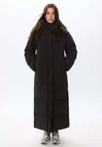 Duga crna puffer jakna s visokim ovratnikom, kopčama na dugmad i kvadratnom teksturom, s dugim rukavima i bočnim džepovima. Ispod su srebrne tenisice.