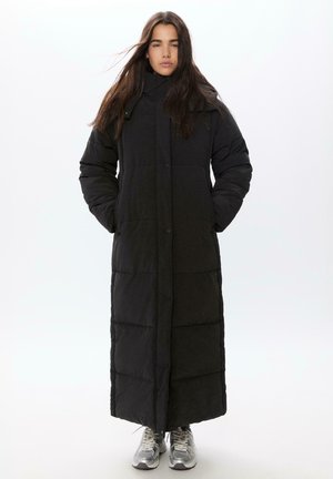 Długi czarny płaszcz puchowy z wysokim kołnierzem, zatrzaskami i pikowanym wzorem, z długimi rękawami i bocznymi kieszeniami. Pod nim srebrne sneakersy.