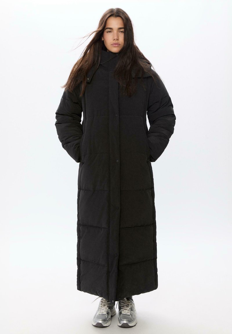 Duga crna puffer jakna s visokim ovratnikom, kopčama na dugmad i kvadratnom teksturom, s dugim rukavima i bočnim džepovima. Ispod su srebrne tenisice.