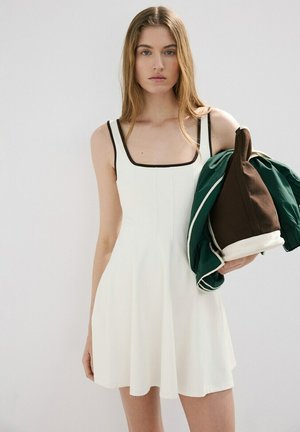 Joven con cabello largo, vestida con un vestido blanco sin mangas con ribete negro, sosteniendo una chaqueta plegada verde y marrón sobre un fondo liso.