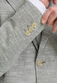 Blazer gris claro con una tela texturizada. Presenta cuatro botones beige en el puño y una camisa blanca visible debajo.
