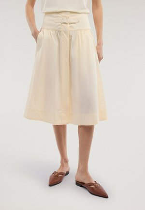 Kobieta w kremowej spódnicy w kształcie litery A z paskiem, z rękami w kieszeniach, w brązowych wsuwanych butach z zaostrzonym noskiem i okrągłymi klamrami.