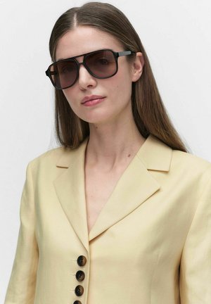 Kobieta z długimi brązowymi włosami, nosząca duże czarne okulary przeciwsłoneczne i jasnżółty żakiet, spoglądająca lekko w bok.