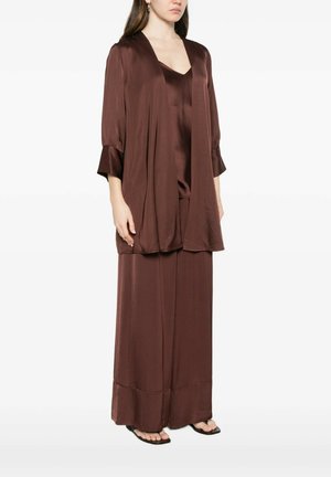 Donna che indossa un completo loungewear largo, setoso, marrone scuro con maniche lunghe, pantaloni a gamba larga e sandali neri con punta quadrata.